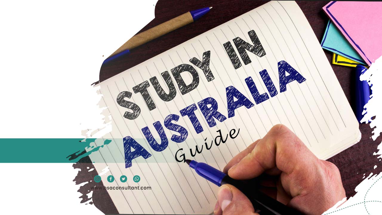 notebook lies down horizontally on the table. A hand is holding a pen & "Study in Australia Guide" is written on notebook